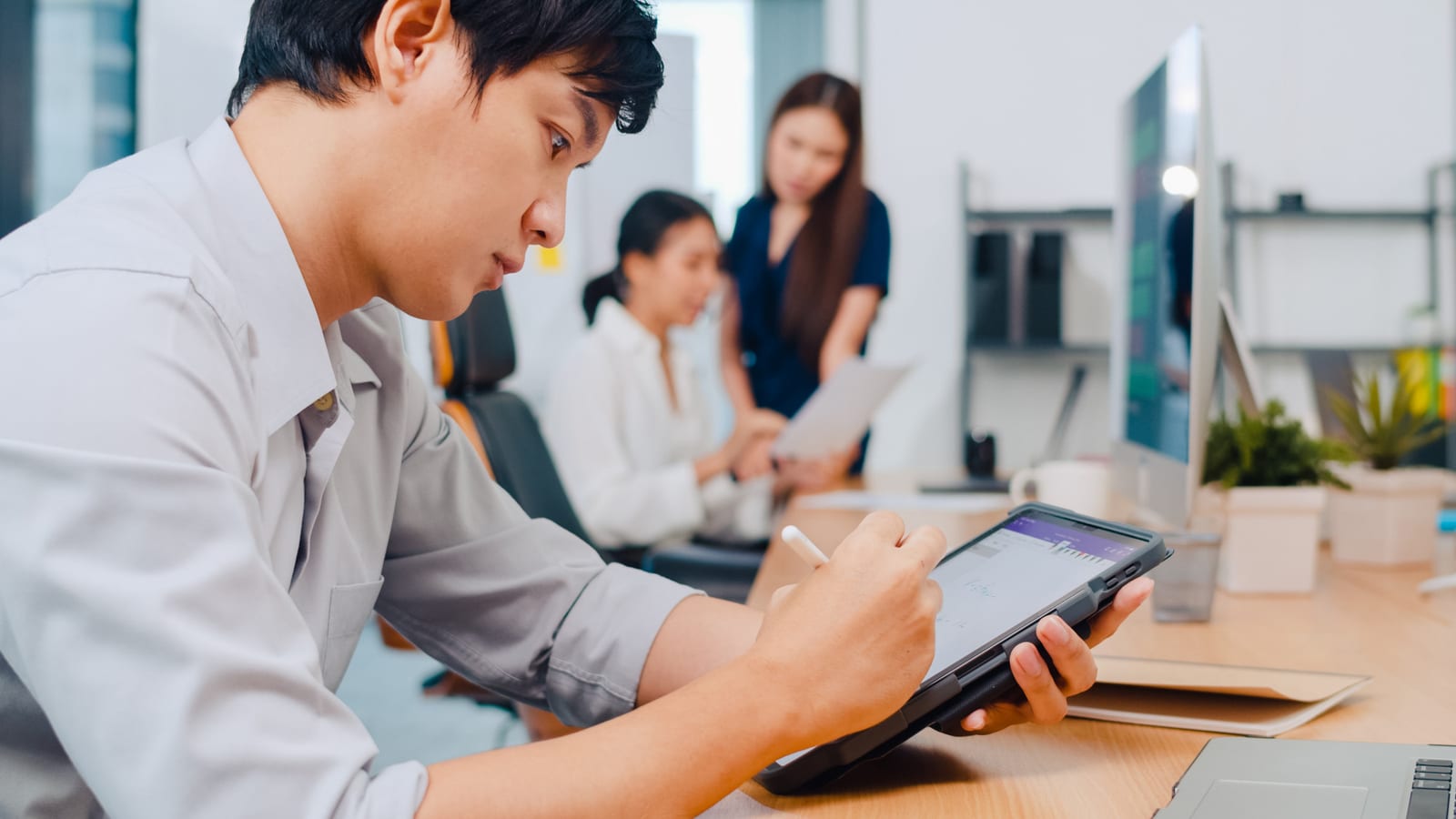 資料を確認する同僚の背後でタブレットに書き込むビジネスパーソン
